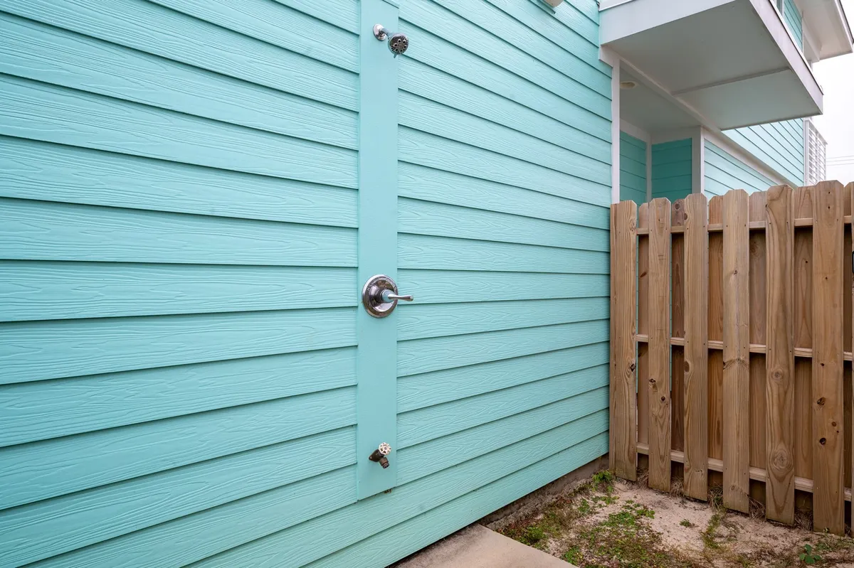 A playful outdoor corner featuring a turquoise wall, wooden fence, and visible outdoor shower. The scene suggests a fun, private spot for rinsing off after beach adventures, set in a cozy, pet-friendly yard.
