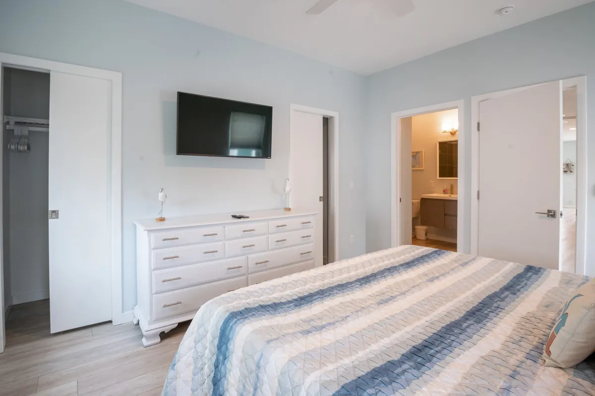 A tranquil bedroom scene featuring a king bed adorned with a patterned quilt, a white dresser, a mounted television, and open doors leading directly to a bathroom. Soft lighting and subtle décor create a restful, private atmosphere ideal for unwinding.
