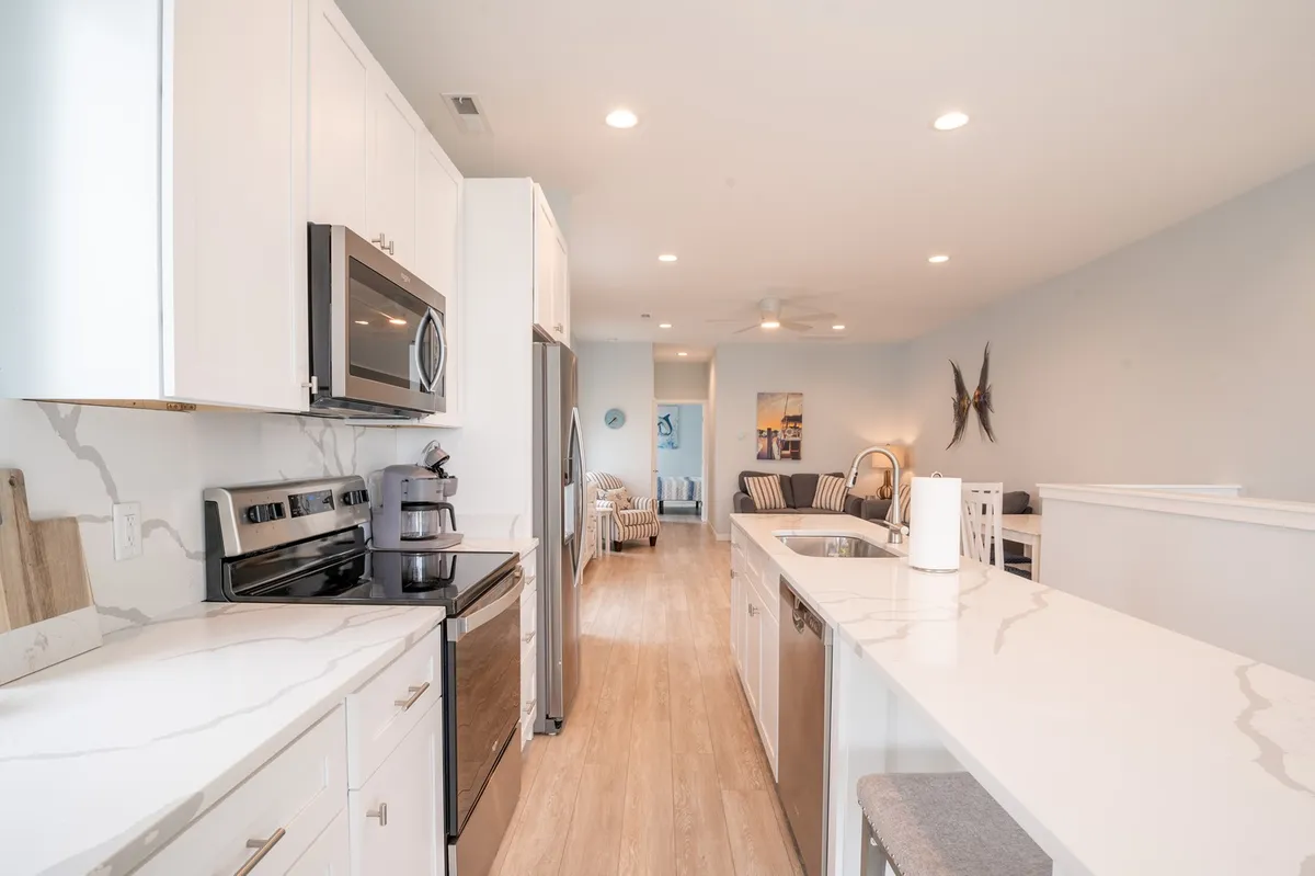 The image captures a welcoming, open-plan kitchen with a sleek quartz island at its center. Stainless steel appliances line the walls, white cabinetry brightens the space, and counter seating invites casual breakfasts or conversation as meals are prepared. The open layout subtly hints at the connection to the dining and living areas, emphasizing the kitchen's social, central role.