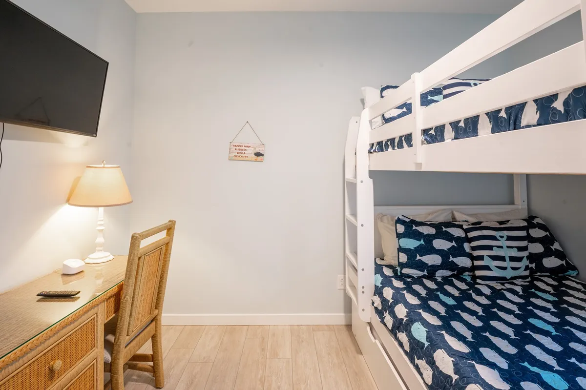 A playful bunkroom featuring a full-over-full bunk bed, wall-mounted TV, desk and chair, table lamp, and decorative sign. The space is inviting for both sleep and play, ideal for kids or groups.