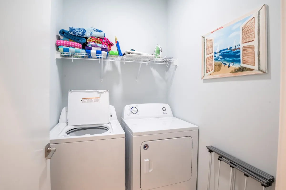 A bright, practical laundry room with a washing machine, dryer, a tidy shelf of colorful towels, and cheerful wall art. The space is functional yet welcoming, making post-beach cleanups easy.