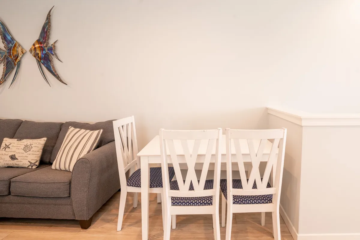 A cozy, light-filled dining and living area is featured, with a gray sofa and a white dining table that seats six. Decorative fish artwork adds coastal charm. The combined spaces are designed for both relaxed dining and comfortable lounging, creating an inviting backdrop for family meals or evening entertainment.