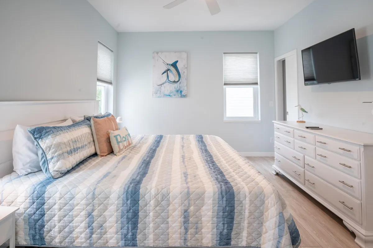 A tranquil bedroom with a striped bedspread, artwork on the wall, a television, and a dresser. The room is bathed in natural light, evoking a sense of calm and comfort—perfect for restful nights after beach adventures.