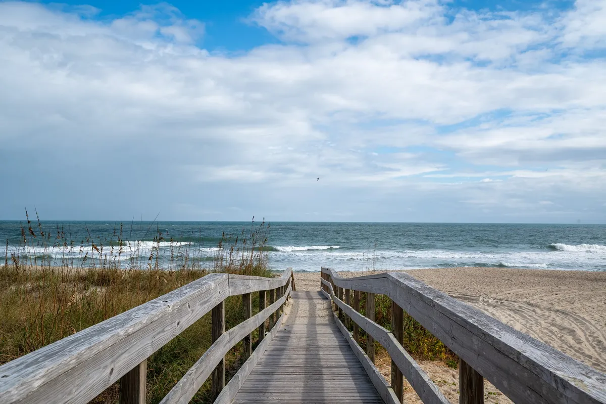 This image shows a wooden boardwalk stretching out towards gentle ocean waves, with the distant horizon visible. The scene is serene, offering a clear connection between the property and the inviting beach environment just steps away.