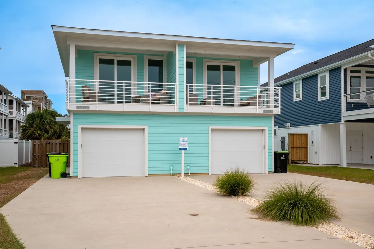 The image showcases the inviting light blue exterior of a spacious condo, featuring large windows that flood the interior with natural light, a welcoming balcony with chairs perfect for soaking in the sunset and ocean breeze, and garage doors for convenient parking. The cheerful atmosphere hints at relaxing mornings with coffee on the balcony, lively family gatherings, and the proximity to Carolina Beach just steps away.